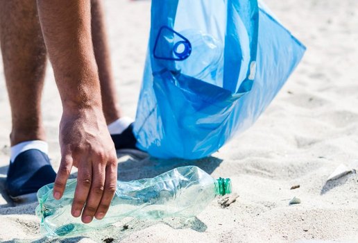 Man hand picking up trash plastic bottle by the beach while holding blue garbage bag