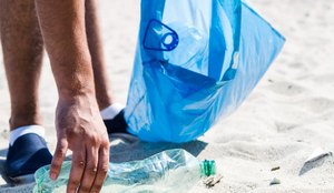 Man hand picking up trash plastic bottle by the beach while holding blue garbage bag